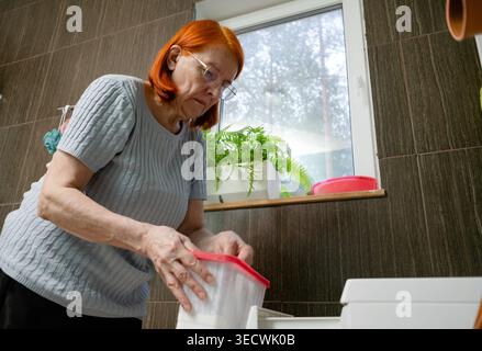 Looking up from a low position, this image captures a senior lady in the act of pouring detergent into her washing machine's compartment. Stock Photo