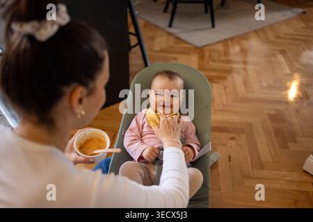 Mom sits on the floor and feeds a small baby with a spoon Stock Photo