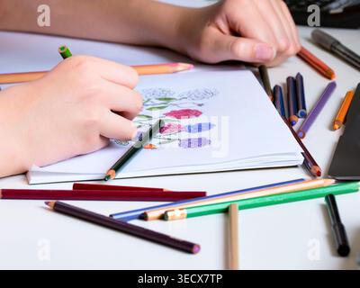 A close-up of a child hands drawing flowers in a sketchbook with various colored pencils on a white desk. Stock Photo