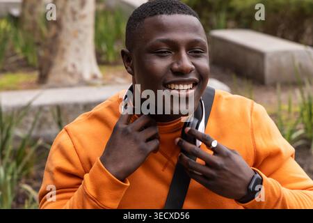 African American adult male sitting on stone bench in park adjusting headphones, in orange crewneck Stock Photo