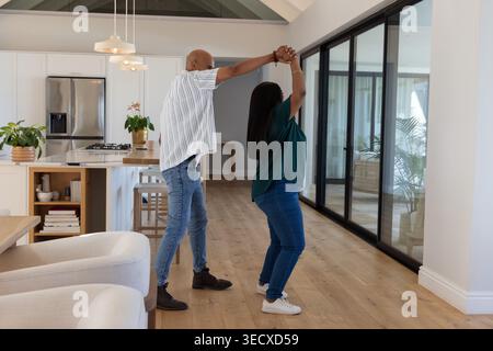 African American middle-aged couple dancing and spinning on hardwood floor near sliding glass doors Stock Photo