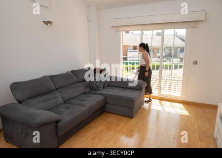 Woman standing beside a large window, looking outside from a home living room Stock Photo