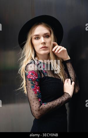 Young blonde woman poses for an outdoor fashion portrait wearing a black wide-brim hat and a black embroidered sheer-sleeve dress in a modern city Stock Photo