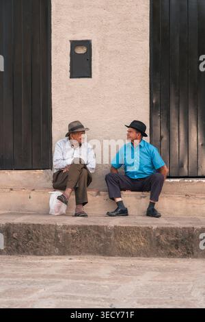Mogotes, colombia - marzo 21, 2026 two older men in hats sit on village steps chatting on a sunny day, sharing relaxed conversation and warm companion Stock Photo