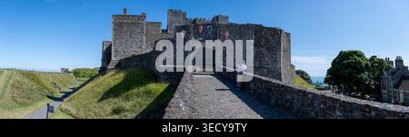 Wide panorama of the stone bridge leading to Dover Castle entrance, with medieval walls, heraldic shields and visitors under a blue sky in Dover, Kent Stock Photo