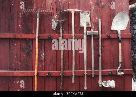 Vintage garden tools hanging on a weathered red wooden wall, with rake, shovel, hoe, pickaxe and fork arranged in rustic farm storage Stock Photo