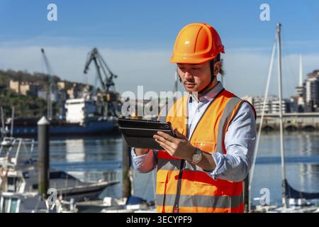 Young asian man wearing a safety helmet and reflective vest, using a digital tablet at a commercial port while managing shipping, cargo, and global lo Stock Photo