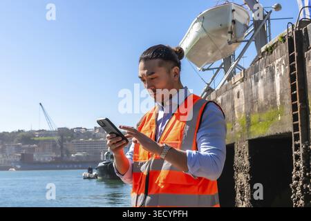 Young asian man wearing a safety vest and using a smartphone outdoors at a cargo port, managing operations and communicating in the maritime shipping Stock Photo