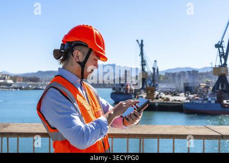 Port worker wearing a hard hat and high visibility vest using a smartphone for managing logistics operations at a cargo terminal with ships and cranes Stock Photo