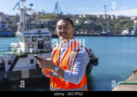 Asian dock worker smiling while standing at a busy port, wearing an orange safety vest and using a smartphone for communication and logistics manageme Stock Photo