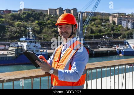 Young man wearing a hard hat and safety vest smiling while using a digital tablet at a busy commercial harbor, managing shipping and transportation lo Stock Photo