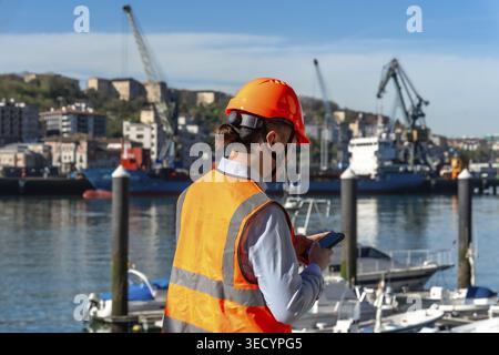 Port worker wearing a safety vest and hard hat using a smartphone for communication and monitoring operations at a busy commercial shipping port with Stock Photo