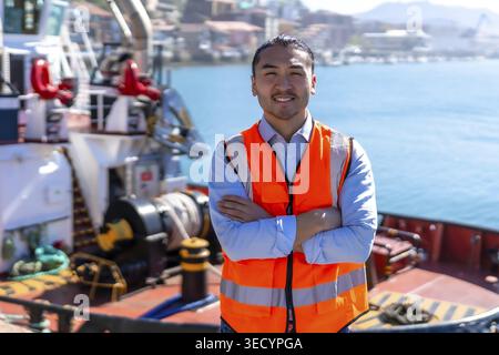 Young asian port worker in orange safety vest stands on a tugboat deck with arms crossed, smiling confidently as harbor cranes and cargo ships form th Stock Photo