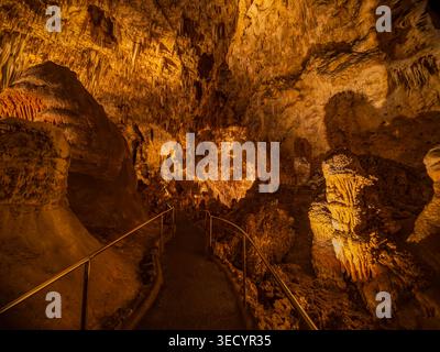 A paved path winds between towering rock formations and stalactites illuminated by warm orange lighting in a deep cavern. Stock Photo