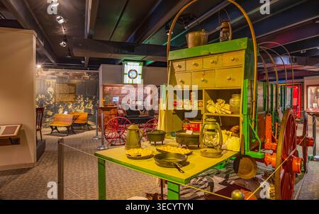 Oklahoma, United States, MAY 24, 2024 - A green vintage farm cart sits inside a museum featuring an open table with cast iron cookware, canned goods, Stock Photo