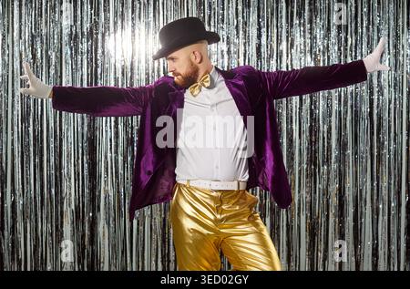 Standing and posing, funny man wearing bowler hat, purple jacket, gold pants sparkly silver backdrop Stock Photo