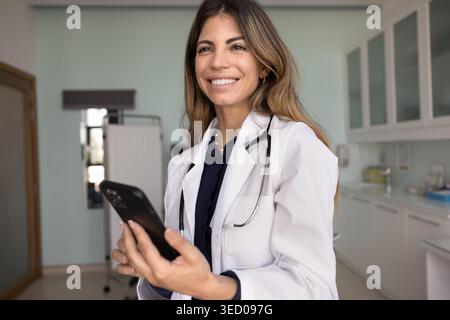 Smiling Latina woman doctor in white lab coat using smartphone Stock Photo