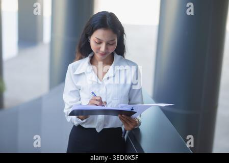 Focused businesswoman writes notes on a clipboard while reviewing charts in a modern office corridor, representing financial analysis, planning, corpo Stock Photo