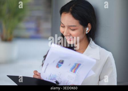 Happy businesswoman with wireless earbuds smiles while reviewing a summary report on a clipboard in a modern office, representing success, communicati Stock Photo