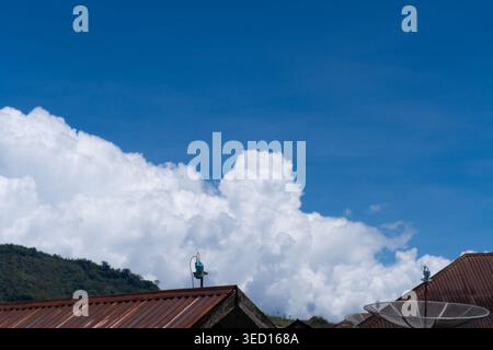 A bright daytime shot of massive white cumulus clouds rising behind traditional house roofs with a satellite dish and green hills in the background. T Stock Photo