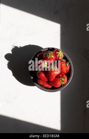 Fresh red strawberries in black bowl on white marble surface. Sunlight casting dramatic shadows. Healthy summer fruit and natural food concept for recipes and nutrition. Stock Photo