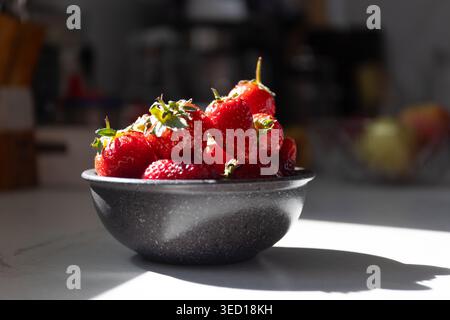 Fresh red strawberries in dark bowl on kitchen counter. Sunlight casting shadows on berries with green leaves. Natural light food scene for healthy eating and summer fruit concepts. Stock Photo