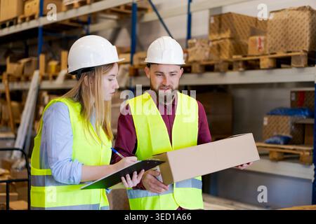 Engaged male and female warehouse workers checking package details on clipboard in distribution center with wooden pallets. Wholesale inventory management and bulk shipping coordination. Systematic stock verification and efficient supply chain documentatio Stock Photo