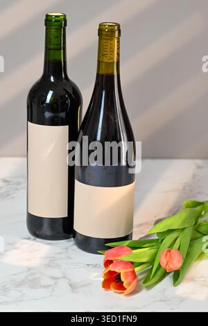 Two wine bottles rest on a marble table next to red tulips. Stock Photo