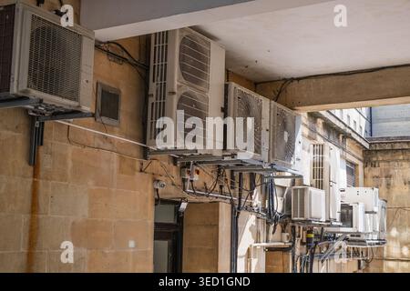 Many air conditioning units, pipes, and wires are densely mounted on a sunlit stone building wall. Stock Photo