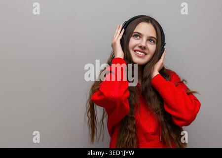 Happy Young Woman Listening To Music. Smiling young woman in a red hoodie enjoys music through headphones, looking cheerful against a simple gray Stock Photo