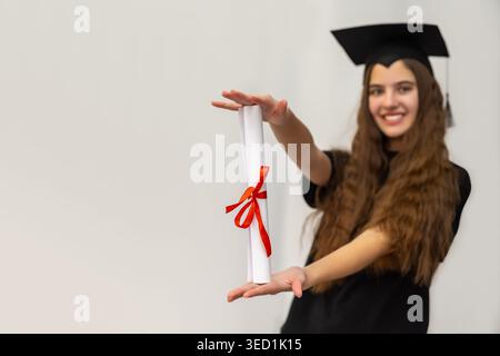Smiling Graduate Holding Diploma Certificate. Young woman in graduation cap proudly presenting a rolled diploma with red ribbon, symbolizing education Stock Photo