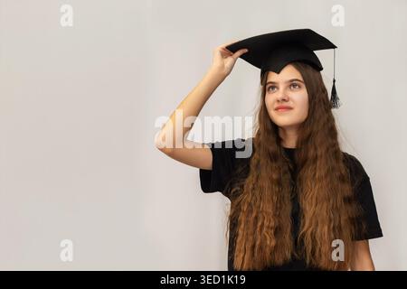 Young Graduate Adjusting Cap With Confidence. Confident young woman wearing a black graduation cap, looking upward with hope and ambition against a Stock Photo