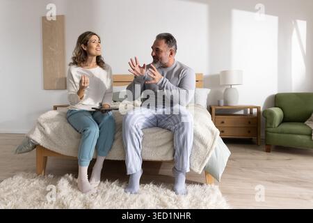 Middle-aged woman and mature man sitting on bed in bedroom, wearing pajamas and holding tablet Stock Photo