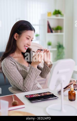 Young Asian woman smiling while applying cosmetics eyeshadow using makeup brush mirror palette at home table beauty Stock Photo
