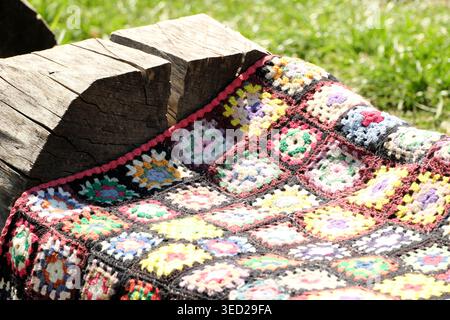 A vibrant crochet blanket rests on a wooden log, basking in warm sunlight. Stock Photo