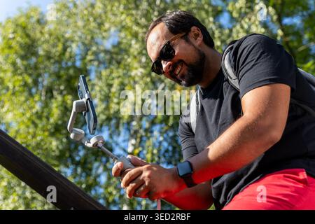 Man uses smartphone gimbal stabilizer filming outdoor video content sunny park green trees. Smiling vlogger create digital media modern equipment Stock Photo
