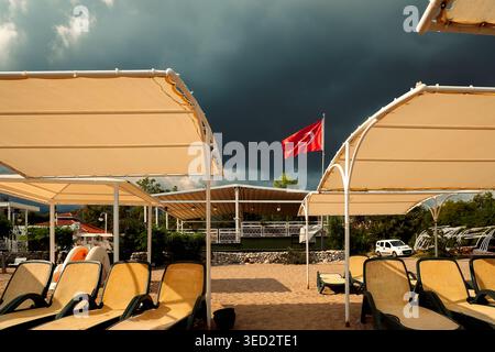 Clouds gather over beach area with sun loungers and Turkish flag flying in wind during late afternoon. dark clouds gathering in the sky. signaling an Stock Photo