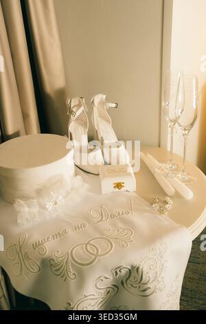 Elegant white heels prepared for  wedding celebration. White high heels sit beside two champagne glasses, small gift box, and decorative item. setting Stock Photo