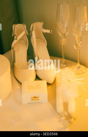 Elegant white heels prepared for  wedding celebration. White high heels sit beside two champagne glasses, small gift box, and decorative item. setting Stock Photo
