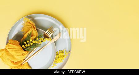 Romantic table setting with mimosa flowers and golden cutlery on yellow background Stock Photo