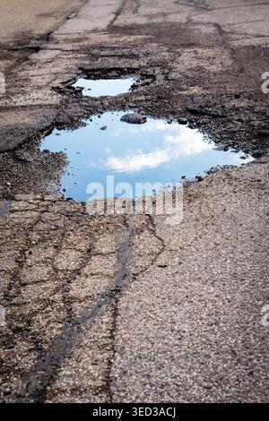 A large, water-filled pothole on a cracked asphalt road reflects a bright blue sky and white clouds, emphasizing road deterioration. Stock Photo