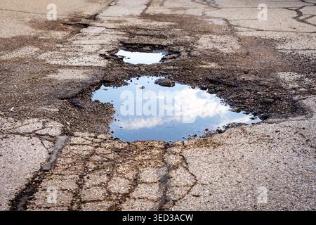 A large, water-filled pothole on a cracked asphalt road reflects a bright blue sky and white clouds, emphasizing road deterioration. Stock Photo