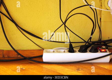 White power strip with four sockets on reddish wooden floor, one plug connected and two black adapters nearby under warm indoor lighting with soft sha Stock Photo