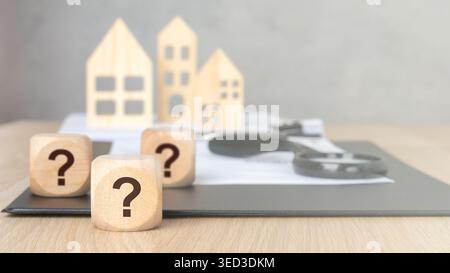 Wooden cubes with printed question marks on a clipboard with documents, a magnifying glass and blurred wooden house models in the background. Concept Stock Photo