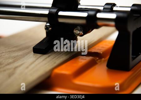 Close-up of an orange mechanical tile cutter, which fixed ceramic tiles of wooden colors, above it is a cutting disc on a white background. The concep Stock Photo