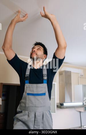 Professional handyman in overalls inspects residential ceiling for repairs and maintenance, looking up with focused attention during home renovation Stock Photo