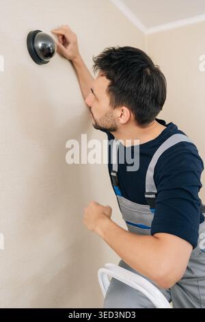Professional handyman in uniform installs home security system. Male technician sets up black dome surveillance camera on wall for safety protection Stock Photo