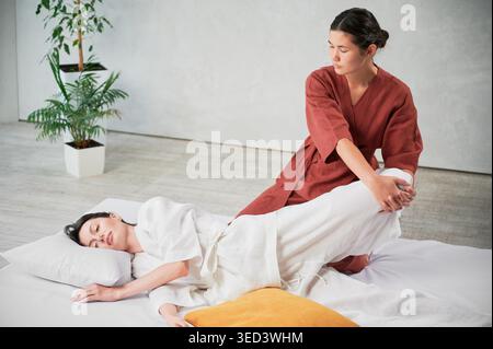 Masseuse making thai yoga massage. Woman therapist doing traditional massage treatment, stretching female patient's body. Client lying on mat, enjoying hand massage in calm atmosphere. Stock Photo