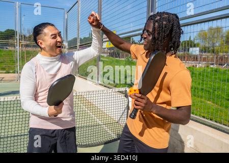 Diverse friends high fiving playing pickleball on outdoor court Stock Photo