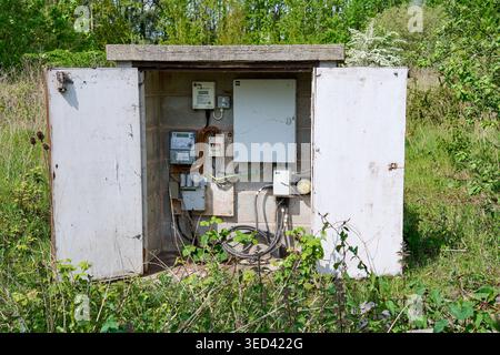 Electrical utility cabinet box standing outdoors with open doors, revealing exposed wiring, meters, and switchboards, surrounded by overgrown grass, r Stock Photo